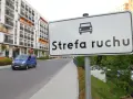 Znak "Strefa ruchu" przed nowoczesnym budynkiem mieszkalnym. Niebieski bus jedzie po drodze.