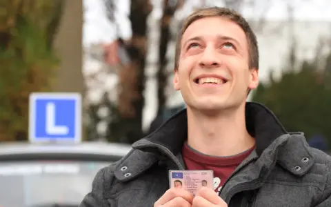 Szczęśliwy młody mężczyzna trzyma prawo jazdy kategorii B, z niebieskim znakiem "L" na dachu samochodu w tle.
