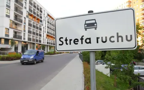 Znak "Strefa ruchu" przed nowoczesnym budynkiem mieszkalnym. Niebieski bus jedzie po drodze.