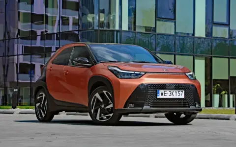 Pomarańczowy Toyota Aygo X Hybrid z czarnymi detalami stoi przed nowoczesnym budynkiem.
