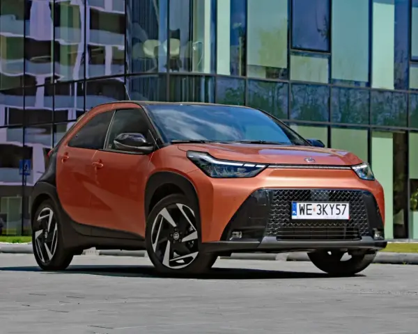 Pomarańczowy Toyota Aygo X Hybrid z czarnymi detalami stoi przed nowoczesnym budynkiem.